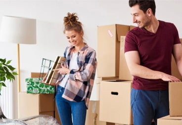 A smiling woman and man unpack cardboard boxes in a bright room, suggesting they are moving into a new home. The woman holds books and a basket, whilst the man carries a box. A lamp and potted plant are visible behind them.