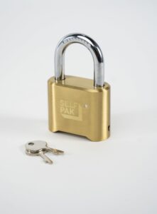 A padlock's combination lock branded "Self Pak" stands upright on a white surface with two silver keys beside it—ideal for those seeking secure, reliable padlocks.