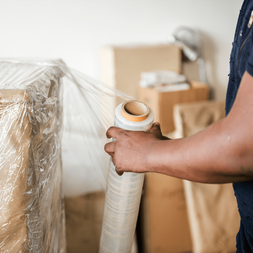 A person wraps a large cardboard box with clear plastic stretch film, with several packed boxes stacked in the background&mdash;an efficient solution often used for self storage or business storage needs.