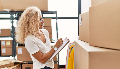 A woman with curly blonde hair holds a clipboard and checks stock in a room filled with large cardboard boxes and metal shelving. Clothing items are visible in the background.