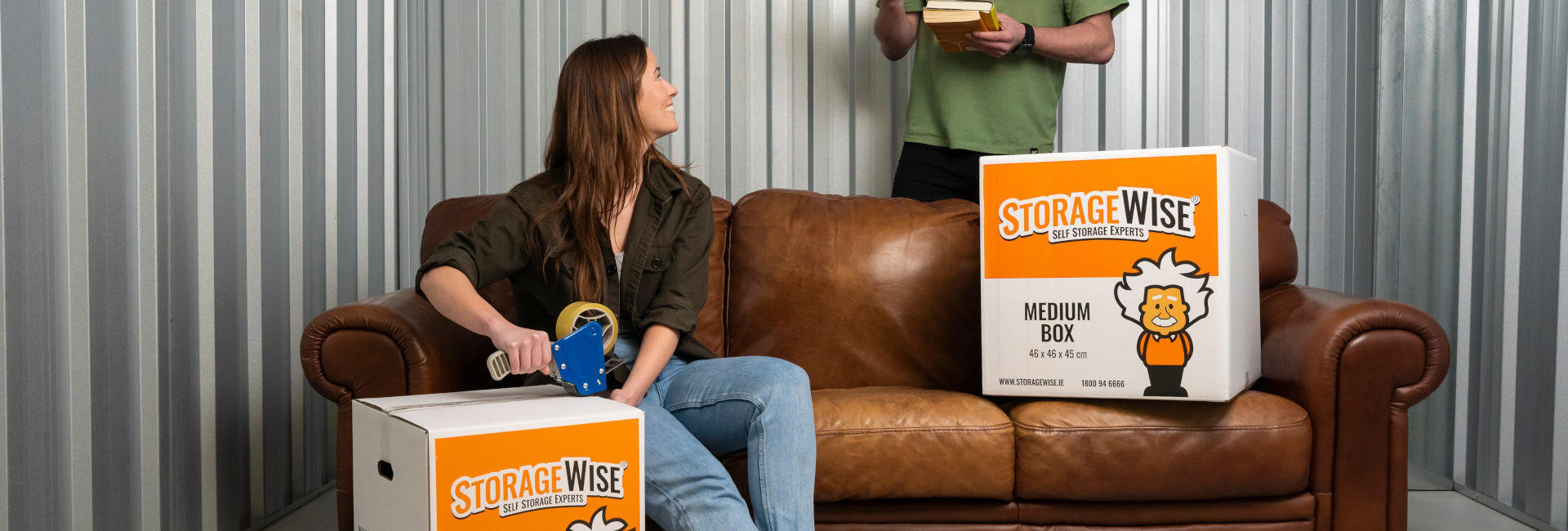A woman sits on a brown sofa sealing a StorageWise moving box for personal self storage while looking up at a man standing beside her holding books, with another StorageWise box on the sofa.