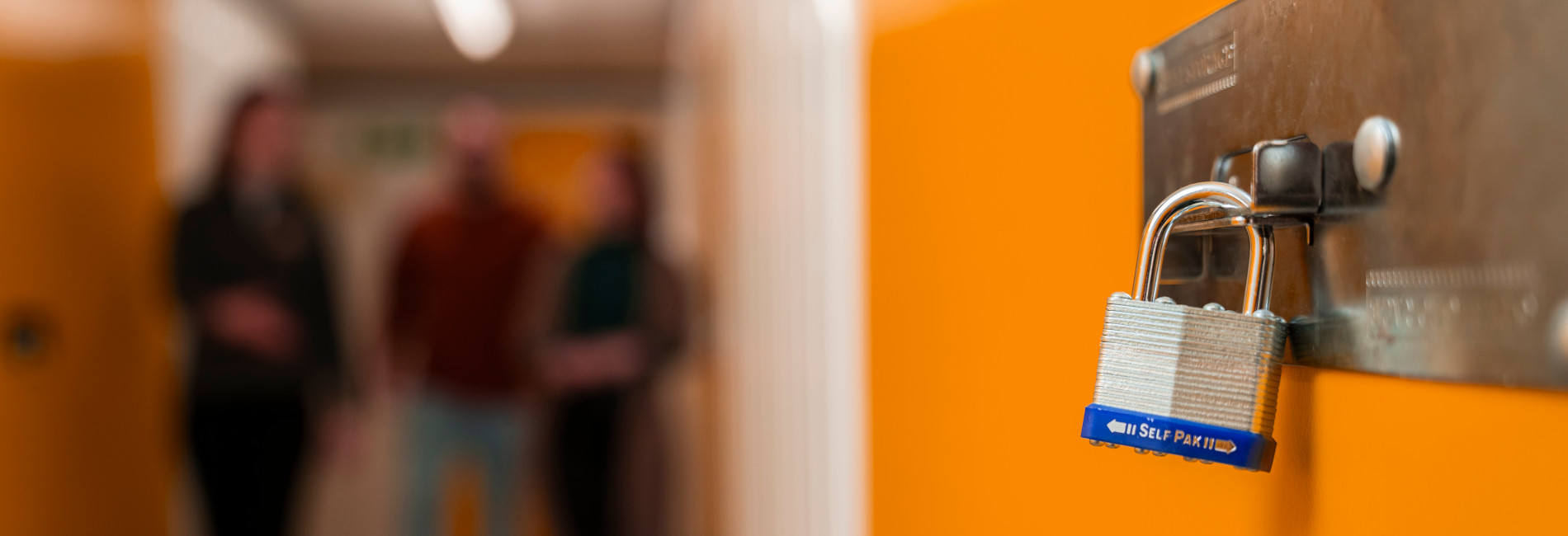A close-up of a silver padlock showcasing security features on a bright yellow door, with three blurred people standing in the background in a corridor.