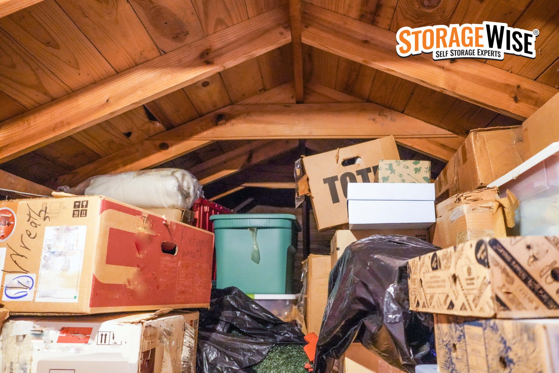 An attic filled with various cardboard boxes, plastic bins, and packed items&mdash;perfect for loft storage solutions. The StorageWise Self Storage Experts logo appears in the top right corner.
