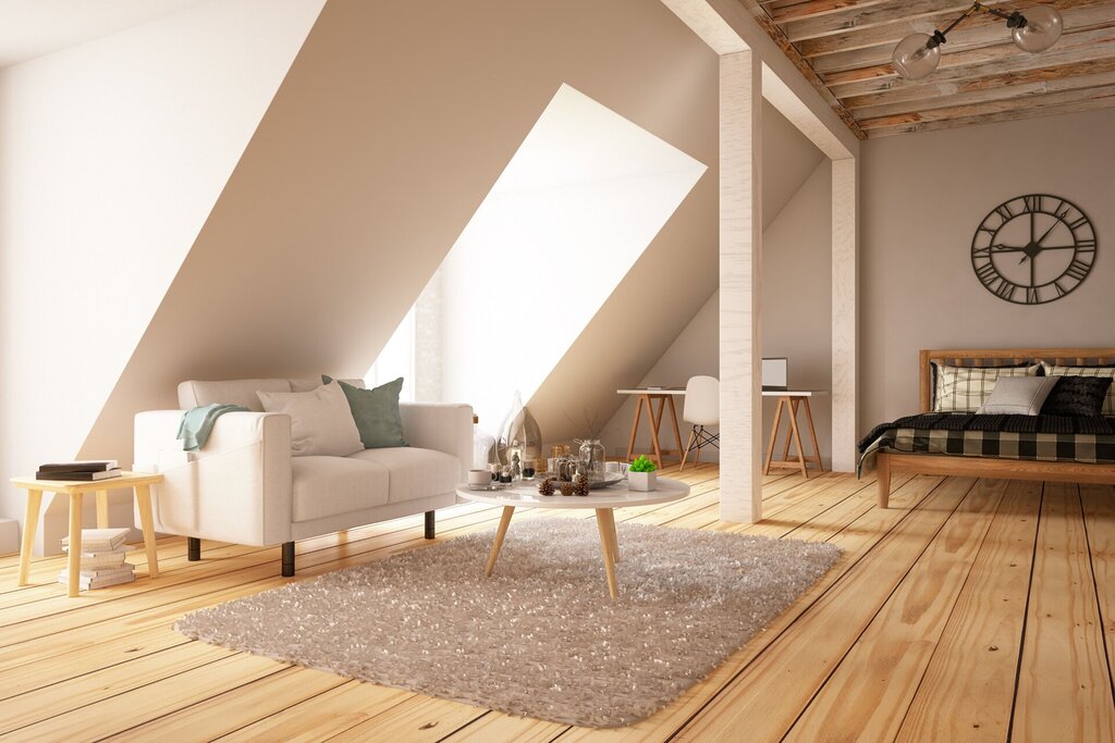 A cosy loft with sloped ceilings features a white sofa, coffee table, and rug in a bright living area. The space also includes a bed, desk, wooden floors, exposed beams, home storage solutions, and a large wall clock.