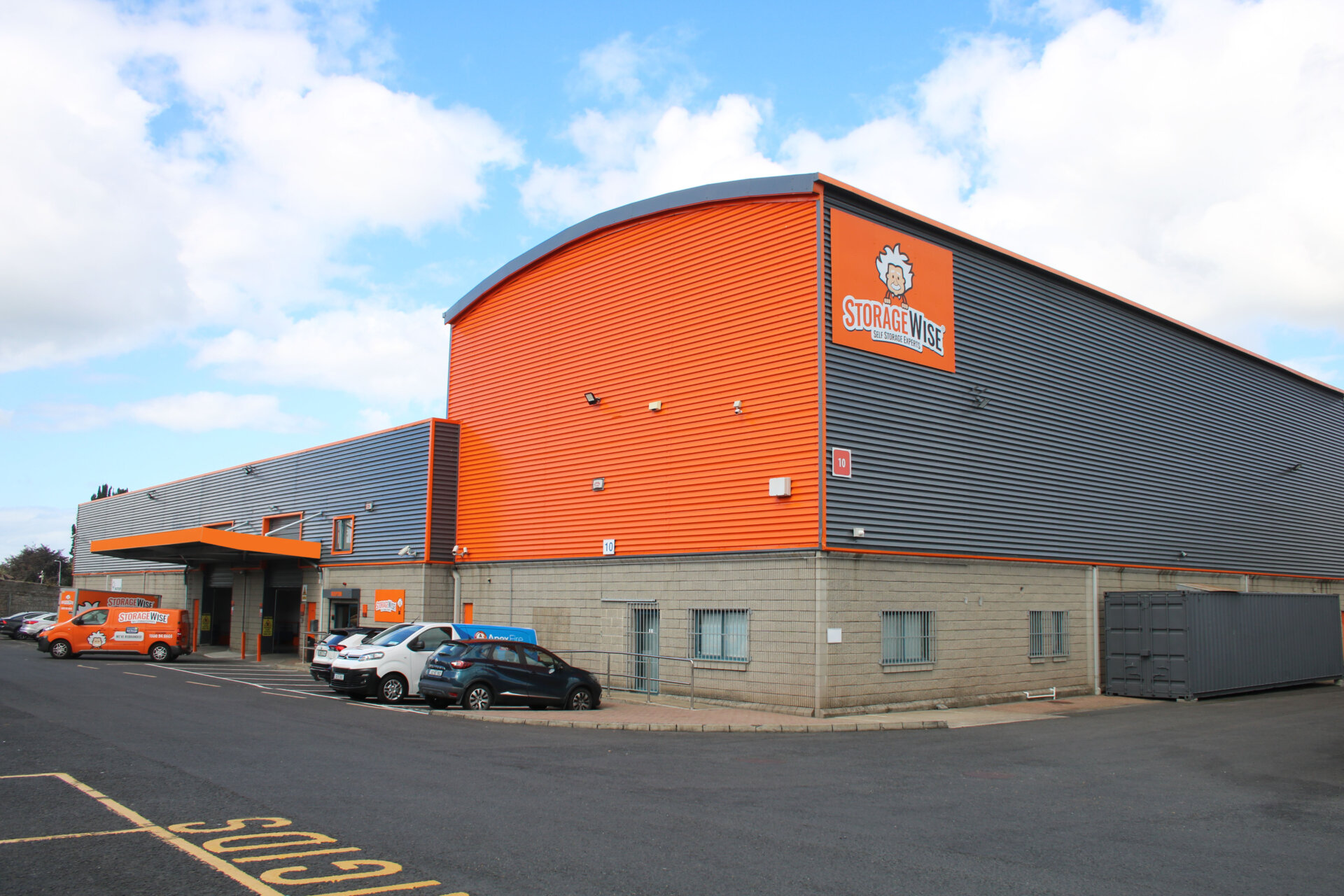 A large, modern self-storage facility with grey and orange cladding, several parked cars, and Storage Wise branding on the building and delivery van offers small businesses a secure way to save money under a partly cloudy sky.