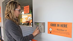 A woman with glasses enters a code on a keypad at the entrance of storage units. A sign beside her reads CODE IN HERE # Number # with instructions for entering the code to access the building.