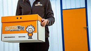 A person in StorageWise uniform holds a StorageWise-branded storage box, ideal for retailers needing extra storage units. The background features a white wall with blue trim and an orange door.