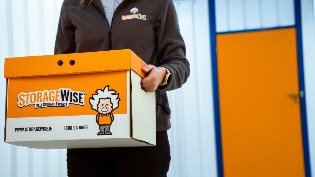 A person in StorageWise uniform holds a StorageWise-branded storage box, ideal for retailers needing extra storage units. The background features a white wall with blue trim and an orange door.