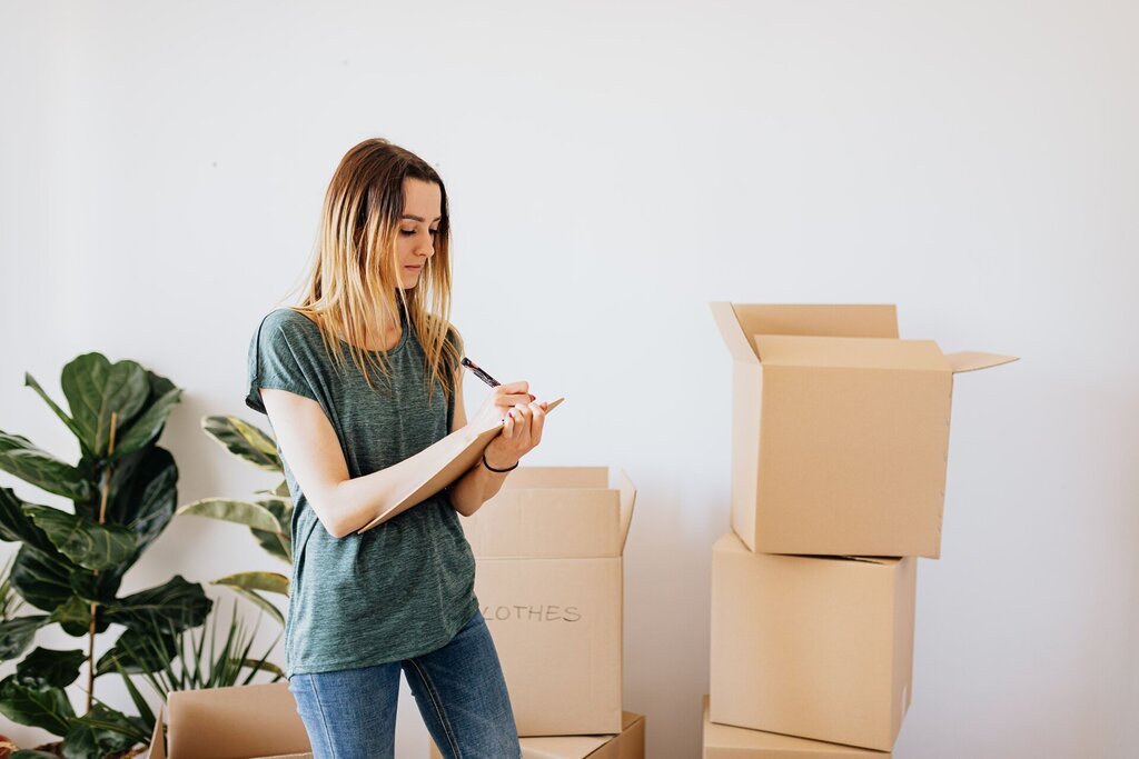 The Benefits of Self Storage While Moving Abroad 2 A woman stands near stacked cardboard boxes, writing on a notepad—perhaps planning for moving abroad or exploring self storage options. She wears a green T-shirt and jeans, with a large potted plant beside her against a white wall.