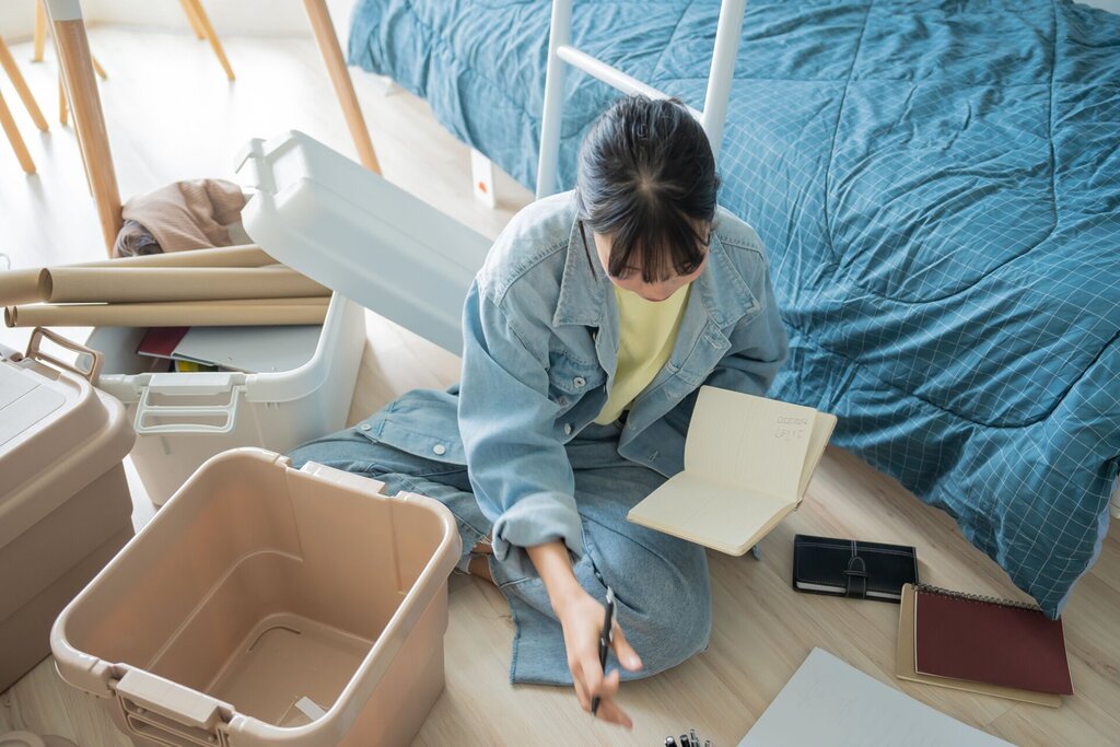 A person sits on the floor next to an unmade bed, surrounded by student storage boxes, notebooks, and papers, holding an open notebook and writing with a pen.