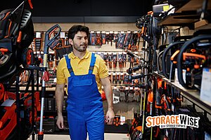 Storing Tools & Equipment: A Guide for Contractors and Trades 1 A man in blue overalls and a yellow shirt walks through an aisle of a storage unit surrounded by storage equipment and tools, perfect for contractors, with the StorageWise logo in the corner.