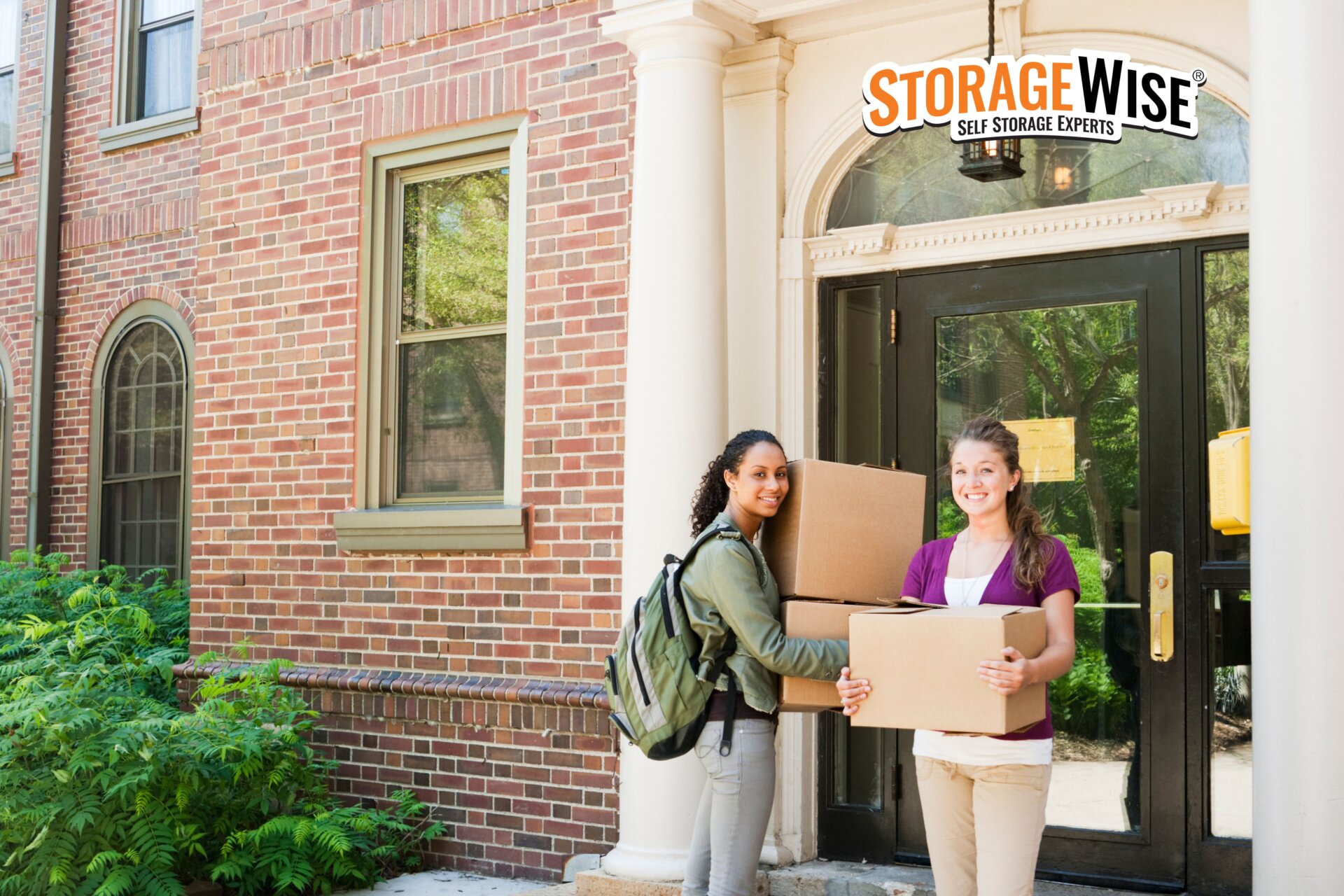 Two women stand outside a brick block of flats, smiling and holding moving boxes near the entrance. Perfect for students in Ireland, use our student packing checklist - Storage Wise Self Storage Experts logo appears in the upper right corner.