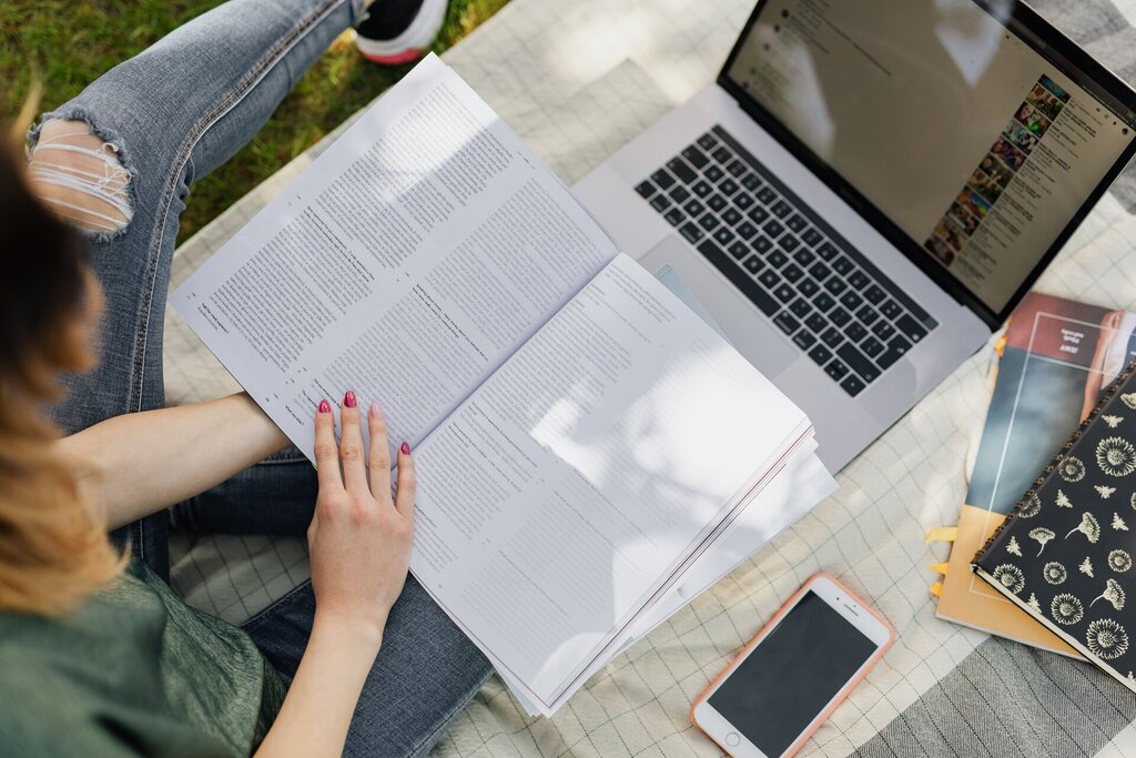 The Ultimate Student Packing Checklist for University in Ireland 1 A person sits on a blanket outdoors, reading an open book with a laptop, phone, and notebooks nearby, suggesting a university Ireland student packing for study or work in a relaxed setting.