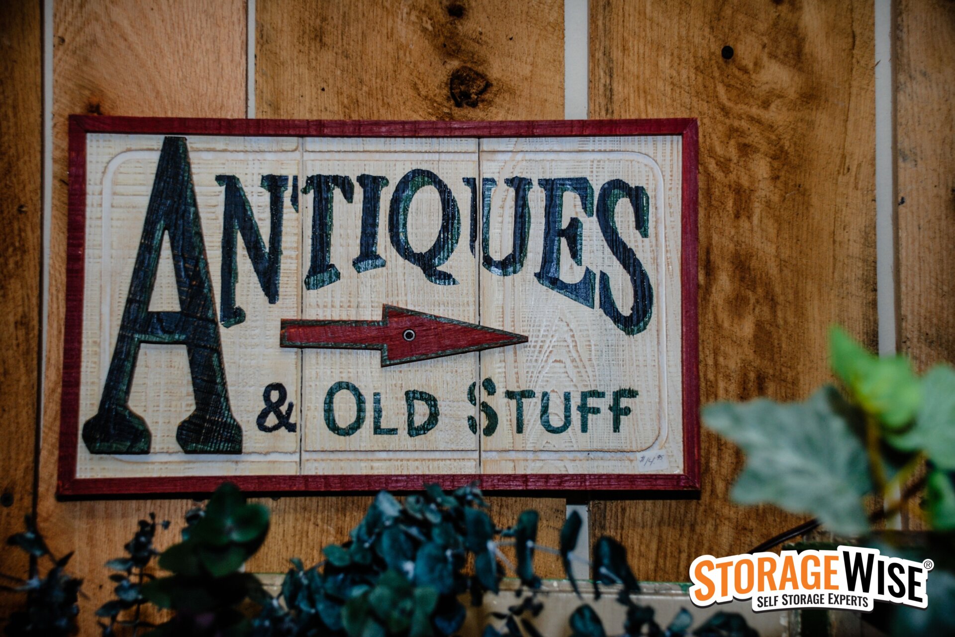 A wooden sign reads ANTIQUES & OLD STUFF with a red arrow pointing right, mounted on a wooden wall. Green leaves are in the foreground, and a StorageWise logo appears in the bottom right corner&mdash;perfect for collectables.