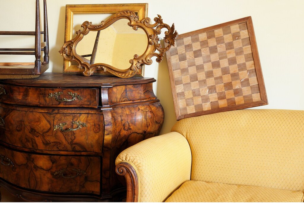A vintage wooden chest of drawers, a decorative gilt-framed mirror, a wooden chessboard, and a yellow upholstered sofa evoke the charm of shop antiques and cherished memorabilia against a pale wall.