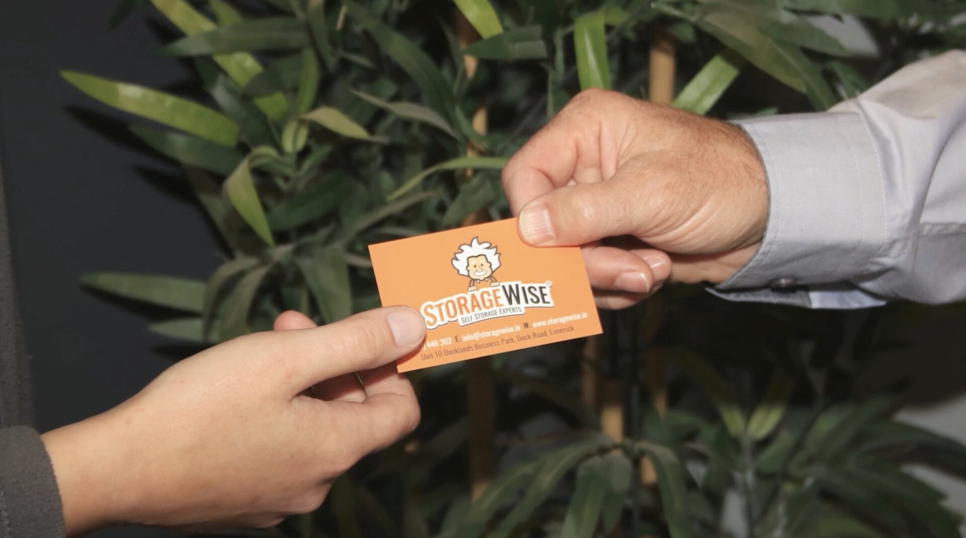 Two people exchange an orange business card that reads Storage Wise with a cartoon logo, in front of leafy green plants&mdash;an ideal networking scene for Irish entrepreneurs seeking dedicated office space in limerick