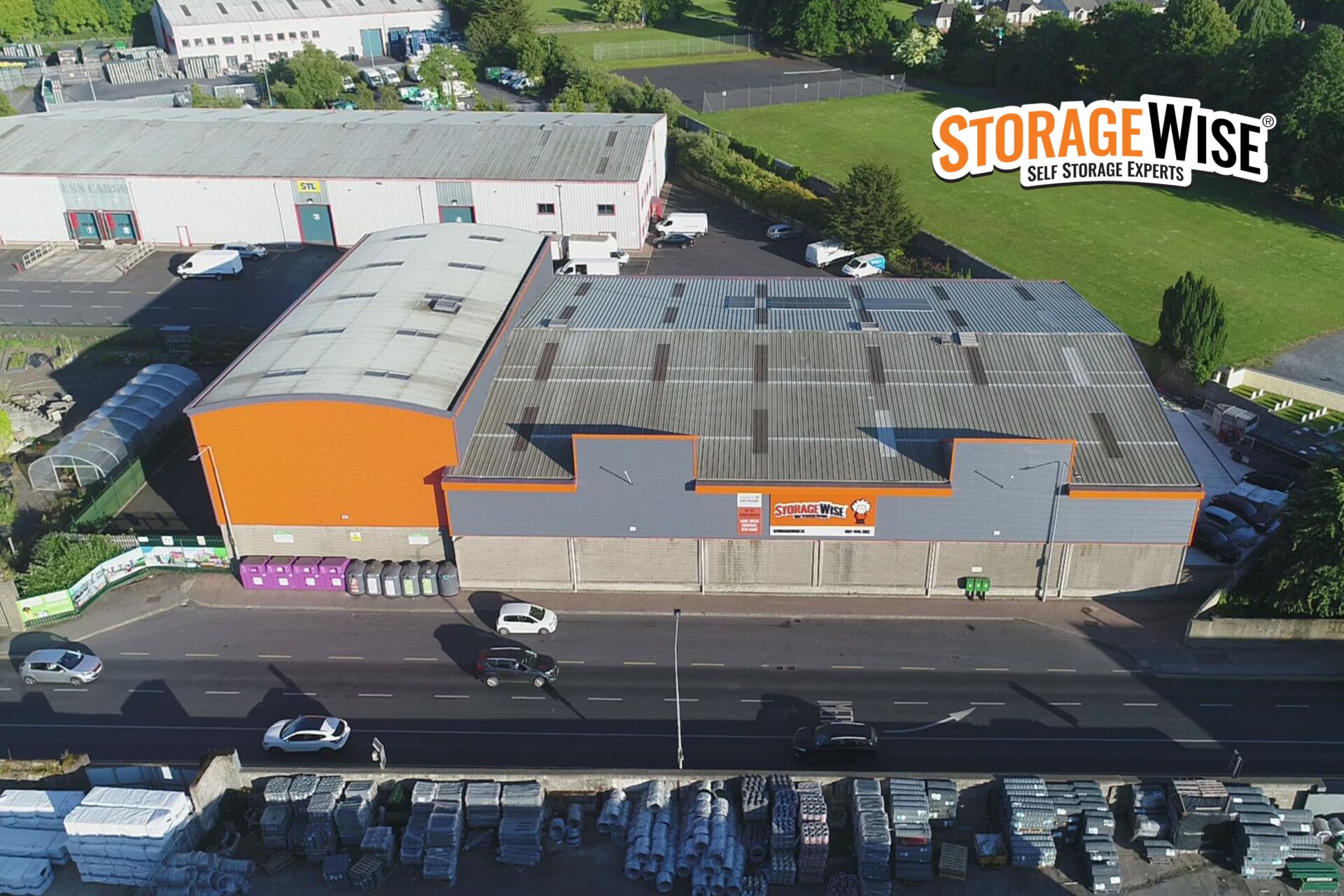 Aerial view of a large, orange and grey self-storage facility next to a road with cars; Storage Wise Self Storage Experts logo in the top right corner. Industrial and green areas surround the building, highlighting convenient storage options for both long-term storage and short-term storage needs.