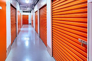 A brightly lit corridor in a self-storage facility with orange roller doors on both sides, clean white walls, and a shiny grey floor, offering flexible storage options for both long-term storage and short-term storage needs.